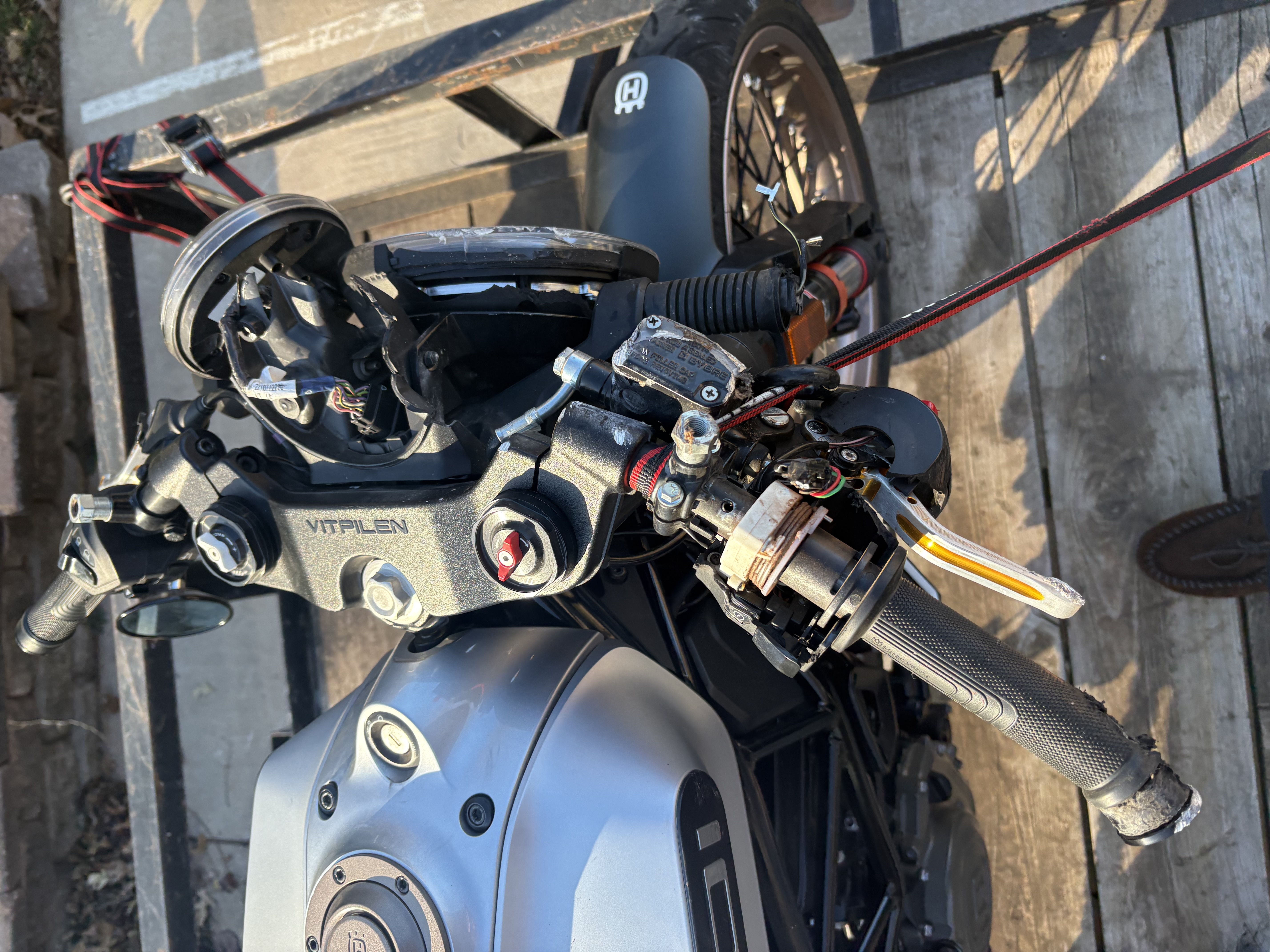 Left handlebar assembly of a Husqvarna Vitpilen motorcycle with exposed wiring, a bent clutch lever and detached headlight housing after a crash; tank and front fender positioned on a wooden trailer outdoors; visible text VITPILEN on the top triple clamp and Husqvarna logo on the fender; tone utilitarian and damaged for mechanical assessment
